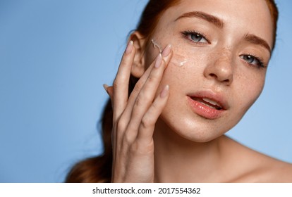 Skin Care. Close Up Of Young Redhead Woman Touching Her Face To Apply Moisturizer, Blue Background