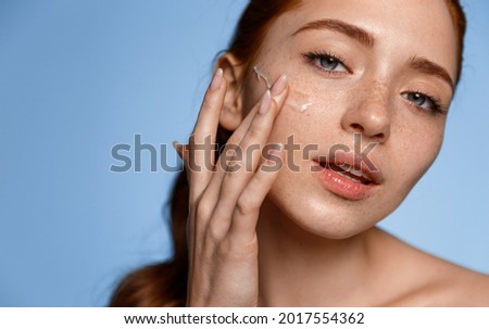 Skin care. Close up of young redhead woman touching her face to apply moisturizer, blue background