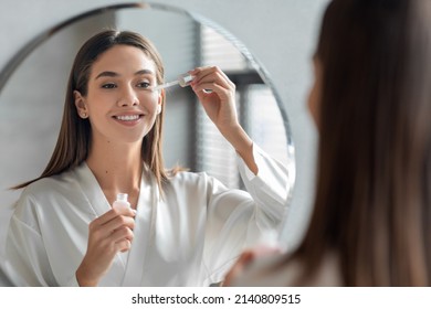 Skin Care Concept. Beautiful Millennial Woman Applying Face Serum Near Mirror, Happy Young Female Standing In Bathroom, Making Daily Skincare Routine At Home, Selective Focus On Reflection