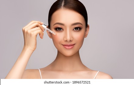 Skin Care Concept. Beauty Portrait Of Chinese Smiling Young Woman Holding Pipette With Cosmetic Oil  Or  Serum Near Clean Face. Beautiful  Asian Girl. Cosmetology And Spa