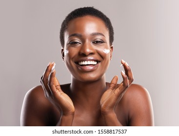 Skin Care Concept. Closeup Portrait Of Joyful African American Woman Applying Hydration Cream On Face, Grey Studio Background