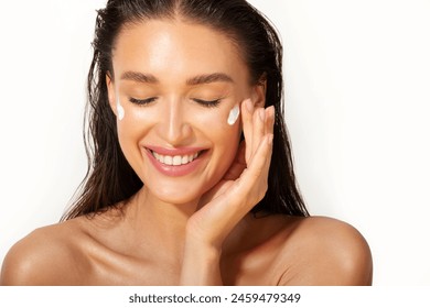 Skin care concept. Happy European woman applying cosmetic moisturizing cream on her face and smiling with closed eyes on white background - Powered by Shutterstock