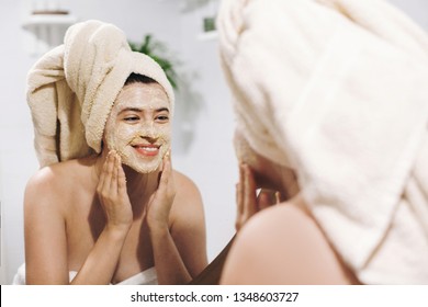 Skin Care Concept. Young Happy Woman In Towel Making Facial Massage With Organic Face Scrub And Looking At Mirror In Stylish Bathroom. Girl Applying Scrub Cream, Peeling And Cleaning Skin