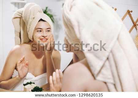 Skin Care concept. Young happy woman in towel making facial massage with organic face scrub and looking at mirror in stylish bathroom. Girl applying scrub cream, peeling and cleaning skin