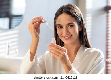 Skin Care Cosmetics. Beautiful Female Holding Bottle With Face Serum And Smiling At Camera, Attractive Young Lady Posing In Bathroom, Enjoying Making Beauty Treatments At Home, Free Space