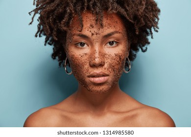 Skin care and cosmetology concept. Beautiful dark skinned serious female model has brown coffee scrub, does peeling of skin, wants to remove wrinkles, poses over blue studio background