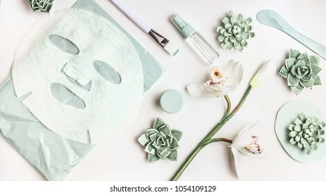 Skin Care Flat Lay With Facial Sheet Mask, Mist Spray Bottle , Succulents And Orchid Flowers On White Desktop Background, Top View. Beauty Spa And Wellness Concept