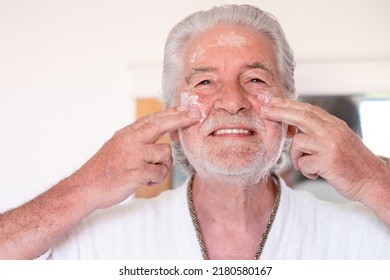 Skin Care. Handsome Elderly Bearded Man In Bathrobe Applying Cream On His Face As A Daily Routine