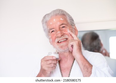 Skin Care. Handsome Elderly Bearded Man In Bathrobe Applying Cream On His Face As A Daily Routine, Holding A Jar Of Cream