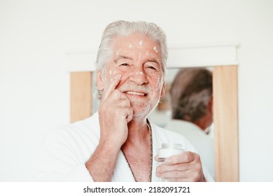 Skin Care. Handsome Elderly Bearded Man In Bathrobe Applying Cream On Face As Daily Routine, Holding A Jar Of Cream