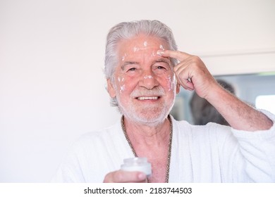 Skin Care. Handsome Elderly Bearded Man In Bathrobe Applying Cream On Face As Daily Routine, Holding A Jar Of Cream