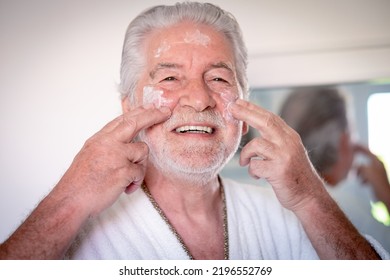 Skin Care. Handsome Elderly Bearded Man In Bathrobe Applying Cream On His Face As A Daily Routine