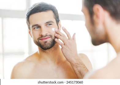 Skin care. Handsome young shirtless man applying cream at his face and looking at himself with smile while standing in front of the mirror 