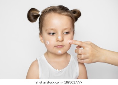 Skin Care, Hygiene, Protection Of Baby's Skin Concept. Close Up Studio Shot Of Mother's Hand, Applying Lotion Cream On Face Of The Little Beautiful Baby Girl After Bathing To Protect Skin.