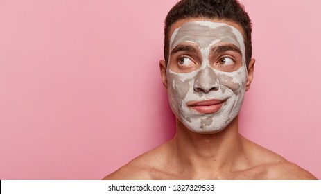 Skin care and peeling concept. Attentive man focused away, has white clay mask, uses beauty products and cosmetics, models over pink background with black space, being model for cosmetologist