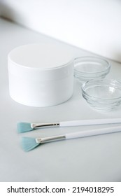 Skin Care Products At Beauty Salon Mockup. Closeup Shot Of White Plastic Jar With Face Cream, Glass Bowl And Peeling Brushes On Grey. Vertical Photo