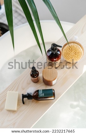 skin care products, natural cream in jar, body lotion in brown bottle, organic soap and massage peeling brush on bamboo caddy and bathtub with bubbles in bathroom, above view