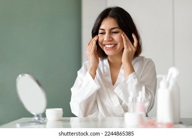 Skin care routine. Happy attractive young middle eastern woman in white silk bathrobe using eyes cream at home, beautiful lady test newest organic beauty product, sitting at vanity table, copy space - Powered by Shutterstock