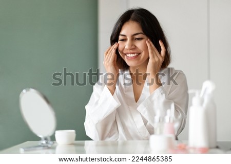 Skin care routine. Happy attractive young middle eastern woman in white silk bathrobe using eyes cream at home, beautiful lady test newest organic beauty product, sitting at vanity table, copy space