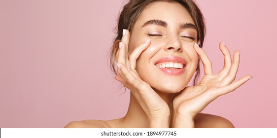 Skin care. Woman with beauty face touching healthy facial skin portrait. Beautiful smiling girl model with natural makeup touching glowing hydrated skin on pink background closeup.