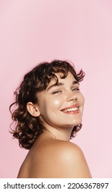 Skin care. Woman with beauty face and healthy facial skin portrait. Beautiful curly girl model with natural makeup touching glowing hydrated skin on pink background closeup. High quality image.