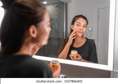 Skin care woman putting face mask product cleaning skincare beauty lifestyle - Asian woman looking in LED mirror in bathroom applying facial cleanser lotion.
