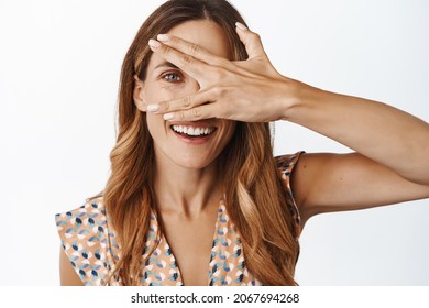 Skin care and women beauty. Portrait of healthy happy middle aged woman, 30 years old, cover face and peeking with one eye at camera, smiling, white background