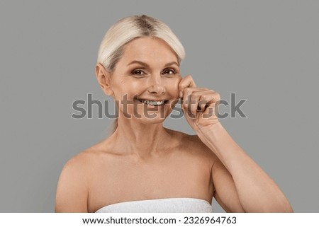 Skin Firming Concept. Beautiful Senior Woman Touching Her Face And Smiling At Camera, Attractive Mature Female With Flawless Tight Skin Standing Wrapped In Towel Over Grey Studio Background