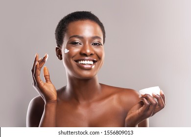 Skin Hydration. Portrait Of Cheerful Black Girl Holding and Applying Moisturizing Cream on Face, Grey Background With Free Space
