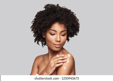 Skin moisturizer. Attractive young African woman applying body cream and smiling while standing against grey background