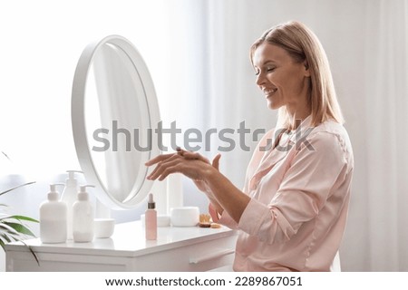 Skin Moisturizing. Smiling Middle Aged Woman Applying Hand Lotion At Home, Beautiful Mature Lady Using Nourishing Serum, Making Daily Skincare Routine While Sitting At Dressing Table In Bedroom