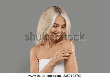 Skin Nourishing. Smiling Beautiful Mature Woman Applying Body Lotion, Portrait Of Attractive Older Lady Wrapped In Towel Using Moisturising Cream After Bath, Posing Over Grey Studio Background