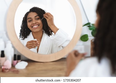 Skin Nutrition. Beautiful Black Woman Applying Organic Moisturizing Serum On Face With Pipette, Making Beauty Treatment At Home, Reflecting In Mirror