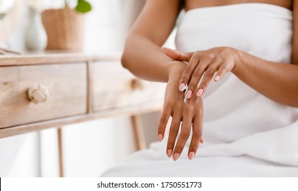 Skin nutrition concept. Unrecognizable black woman applying moisturizing cream to her hands, sitting wrapped in towel in bedroom, cropped image