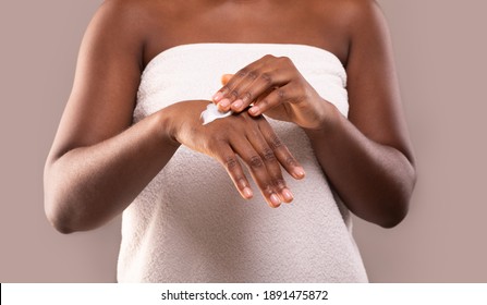 Skin Nutrition. Unrecognizable Black Woman Applying Moisturizing Body Lotion On Hands, Using Rich Nourishing Cream For Skin, African American Lady Standing Wrapped In Bath Towel Over Grey Background