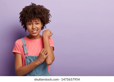 Skin problem concept. Displeased Afro female scratches itchy arm, has allergy, smirks face and looks with dissatisfaction, wears casual pink t shirt and jean overalls, stands against purple wall