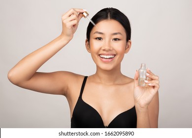 Skincare. Beautiful Asian Girl Applying Cosmetic Oil On Face Skin Standing Over Gray Background. Studio Shot