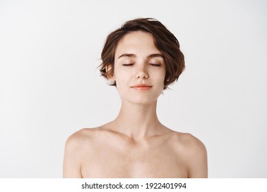 Skincare and beauty. Close-up of young woman with no makeup and naked shoulders, close eyes and smiling tenderly, enjoying fresh and clean feeling after shower, white background.