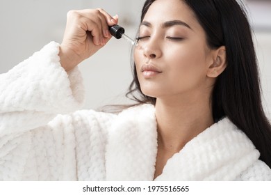 Skincare And Beauty Concept. Portrait Shot Of Young Asian Woman Applying Serum Or Essential Oil On Facial Skin. Model In White Bathrobe Moisturizing Derma With Vitamin E, Collagen And Hyaluronic Acid