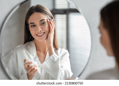 Skincare Concept. Attractive Young Female Looking At Mirror And Applying Moisturising Face Cream, Smiling Millennial Woman Making Beauty Routine At Home, Enjoying Her Skin Condition, Selective Focus - Powered by Shutterstock