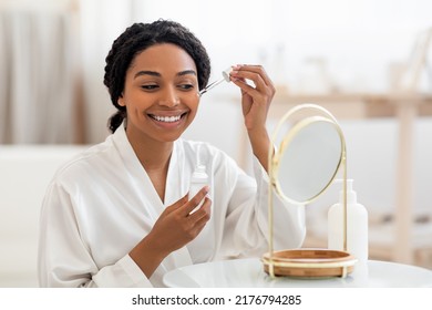 Skincare Concept. Beautiful Black Lady Applying Face Serum With Dropper While Sitting In Front Of Mirror At Home, Smiling African American Woman Moisturizing Skin, Enjoying Self-Care Routine