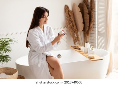 Skincare Concept. Cheerful Brunette Woman Holding Cream Moisturizer Caring For Skin Sitting In Modern Bathroom At Home. Female Beauty Rituals, Skin Care Cosmetic Products Concept - Powered by Shutterstock