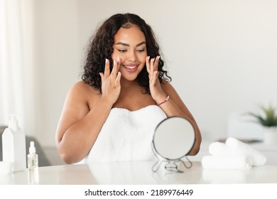 Skincare concept. Happy black oversize woman touching her face while looking at mirror in bedroom, enjoying skin condition after beauty treatments, copy space - Powered by Shutterstock