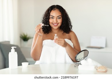 Skincare concept. Plus size black lady applying face serum with dropper while sitting at home, smiling at camera. African american woman moisturizing skin, enjoying self-care routine - Powered by Shutterstock