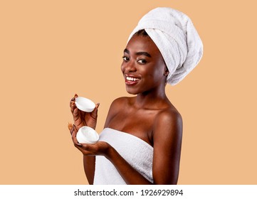 Skincare Concept. Portrait of beautiful young african american woman holding open cream bottle and showing it to camera. Smiling black lady wearing white towel on head isolated on studio background