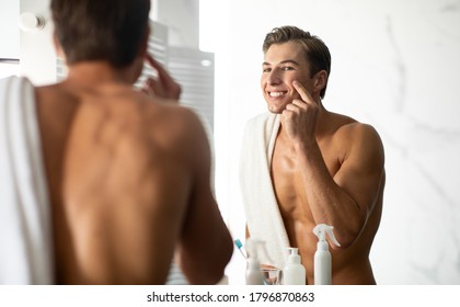 Skincare Concept. Portrait of charming shirtless guy applying cream on face, touching his cheek, looking in mirror