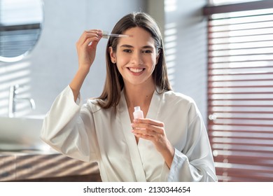 Skincare Concept. Smiling Young Woman Applying Face Serum With Dropper While Making Beauty Routine In Bathroom At Home, Attractive Lady Moisturizing Skin And Smiling At Camera, Copy Space