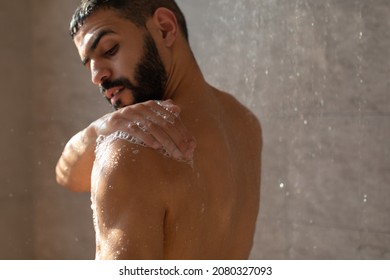 Skincare Concept. Young muscular man taking refreshing shower washing his body, back and bare shoulders with soap foam. Confident handsome guy standing in the bathroom under falling hot water drops