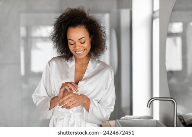 Skincare Cosmetics. Smiling Black Young Woman Applying Moisturizer Cream On Hands, Standing In Bathroom At Home. Lady Enjoying Self Care Routine Moisturizing Skin Indoors - Powered by Shutterstock