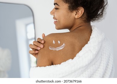 Skincare, dermatology concept. Back view of happy afro american woman with sunscreen cream painted smile on shoulder, applying moisturizing after sun or body lotion at smooth skin, enjoying procedure